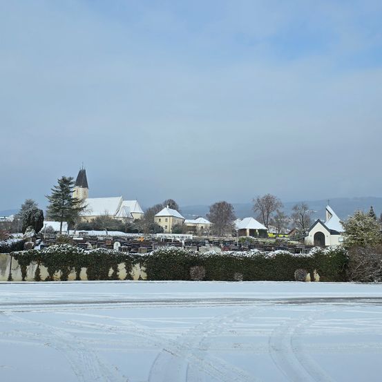 Eine verschneite Landschaft mit einer Kirche, einem Friedhof und entfernten Bergen. Bäume und Büsche sind in Schnee gehüllt.