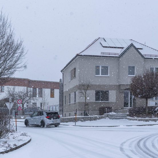 Eine verschneite Straße mit einem abbiegenden Auto, ein Haus mit Solarmodulen auf dem Dach und laublose Bäume.