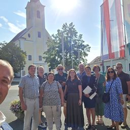 Eine Gruppe von Menschen, darunter Männer und Frauen, steht an einem sonnigen Tag vor einer Kirche. Sie alle lächeln und posieren für ein Foto.
