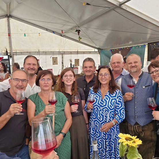Eine Gruppe von Menschen posiert für ein Foto unter einem Zelt, hält Weingläser mit rotem Wein in der Hand, lächelt und schaut in die Kamera.