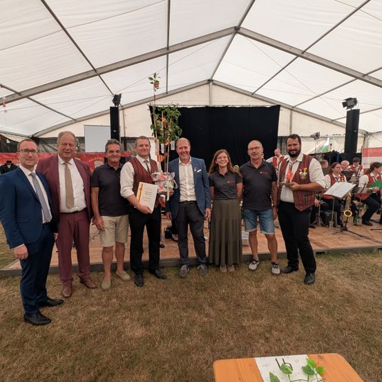 Eine Gruppe von Menschen in formeller Kleidung steht auf einer Bühne unter einem Zelt und hält Auszeichnungen und Zertifikate in den Händen. Im Hintergrund spielt eine Band.