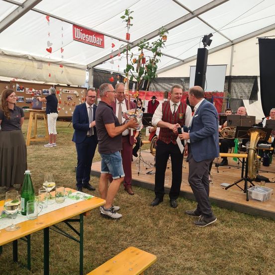 Eine Gruppe von Menschen steht in einem Zelt mit einer Bühne, ein Mann hält einen Blumenstrauß und ein anderer ein Mikrofon. Ein Tisch mit Weingläsern und einer Flasche ist in der Nähe. Eine Band spielt im Hintergrund.