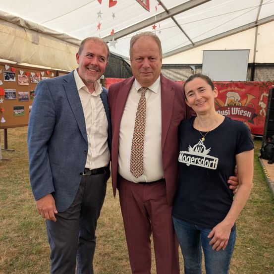 Drei Menschen stehen unter einem Zelt und lächeln für ein Foto. Ein Mann in einem blauen Anzug, ein Mann in einem roten Anzug und eine Frau in einem schwarzen T-Shirt. Hinter ihnen ist ein Banner mit Text und eine Projektionsleinwand.
