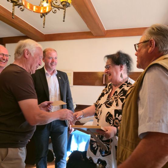 Eine Gruppe von Menschen steht in einem Raum, lächelt und hält Papiere in der Hand. Ein Mann in einem braunen T-Shirt reicht einer Frau in einem Blumenhemd ein Stück Papier.
