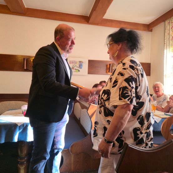 Ein Mann und eine Frau schütteln sich in einem Restaurant die Hände, mit einigen Leuten, die an Tischen dahinter sitzen. Der Mann trägt einen Anzug und die Frau ein Blumenhemd.