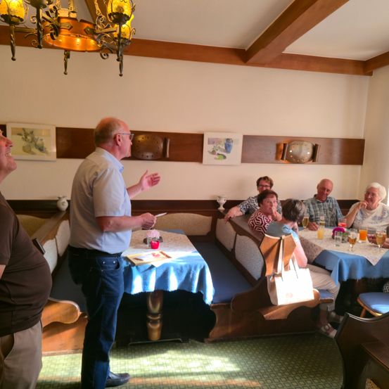 Eine Gruppe älterer Menschen sitzt an einem Tisch in einem Restaurant. Ein Mann steht vor ihnen und spricht, während er gestikuliert. Der Tisch ist mit Getränken und Essen gedeckt.