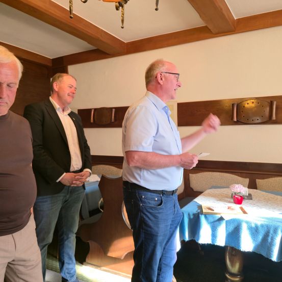 Drei Männer in einem Restaurant, einer hält ein Papier, ein anderer lächelt und der dritte schaut etwas an. Ein Tisch mit einer Blume und einer Kerze ist im Hintergrund.
