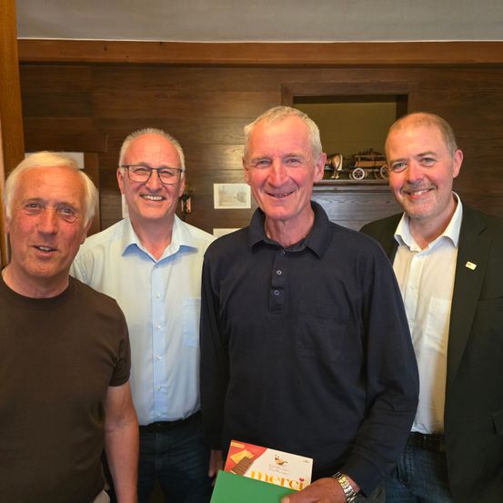 Vier Männer stehen zusammen in einem Raum, lächeln und posieren für ein Foto. Der Mann in der Mitte hält ein Buch mit dem Titel 'Merci'. Hinter ihnen ist eine Holzwand mit einem Bilderrahmen und einem kleinen Spielzeugauto.