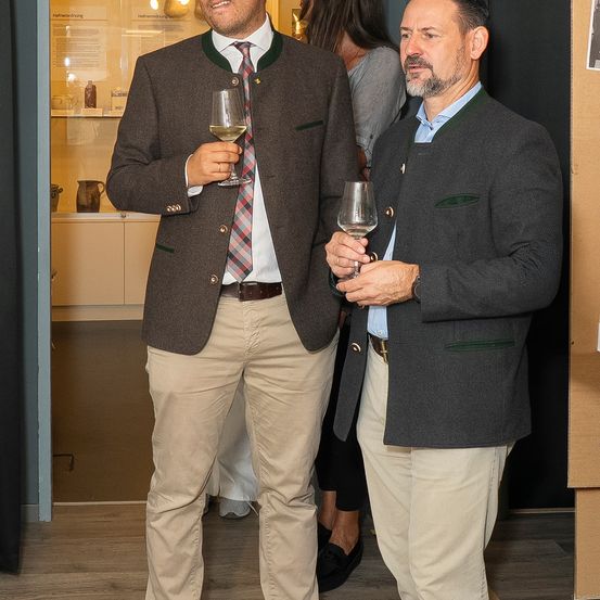 Zwei Männer in traditioneller Kleidung, einer hält ein Weinglas, stehen in einem Raum mit einer Glasvitrine im Hintergrund.
