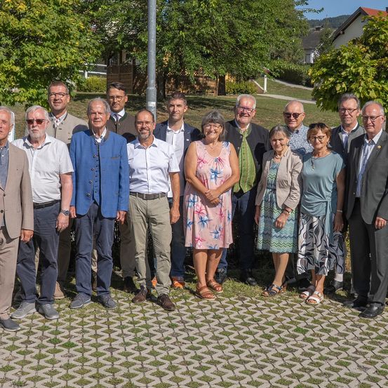 Eine Gruppe von Menschen, sowohl Männer als auch Frauen, in formeller Kleidung, stehen zusammen in einem Park. Sie lächeln und posieren für ein Foto.