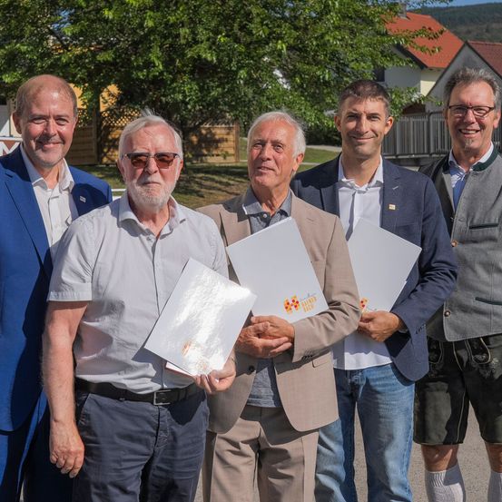 Fünf Männer stehen zusammen und halten Papiere in der Hand, lächeln für ein Foto. Sie sind in verschiedenen Kleidungsstücken gekleidet, einer trägt ein traditionelles deutsches Outfit. Hinter ihnen steht ein Baum und ein Haus mit einem roten Dach.