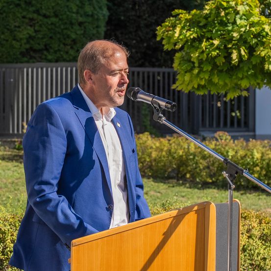 Ein Mann in einem blauen Anzug spricht in ein Mikrofon an einem Rednerpult im Freien.