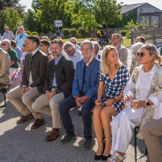 Eine Gruppe von Menschen sitzt in einer Reihe, wahrscheinlich bei einer Veranstaltung im Freien. Sie sind formell gekleidet, einige tragen Brillen und Sonnenbrillen. Hinter ihnen stehen Bäume und ein Gebäude.