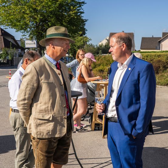 Zwei Männer in formeller Kleidung unterhalten sich auf einer sonnigen Straße. Hinter ihnen sitzt eine Frau mit einer rosa Mütze an einem Tisch. Ein Mann in Hut und traditioneller Kleidung steht in der Nähe.