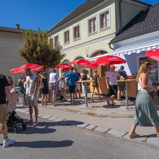 Ein geschäftiges Outdoor-Café mit Menschen, die Getränke unter roten Sonnenschirmen genießen. Eine Frau in einem Rock geht vorbei.