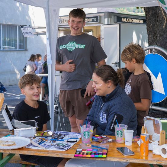 Eine Gruppe von Menschen, darunter eine Frau und drei Kinder, sitzen an einem Tisch mit Kunstmaterialien, lächeln und genießen. Der Tisch ist unter einem Zelt mit einem Schild für Mini-Donuts aufgebaut.