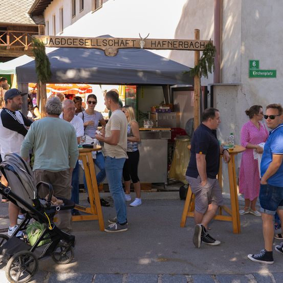 Eine Gruppe von Menschen versammelt sich um einen Essensstand mit einem Vordach, beschriftet mit 'Jagdgesell schaft Hafnerbach.' Sie stehen und sitzen an Tischen, einige mit Getränken und Essen. Ein Kinderwagen steht in der Nähe, und ein Schild an der Wand sagt 'Küchenplatz.'