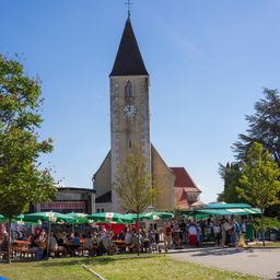Ein sonniger Tag auf einem Dorfplatz mit einem Kirchturm im Hintergrund und Leuten, die unter Schirmen an Tischen sitzen.