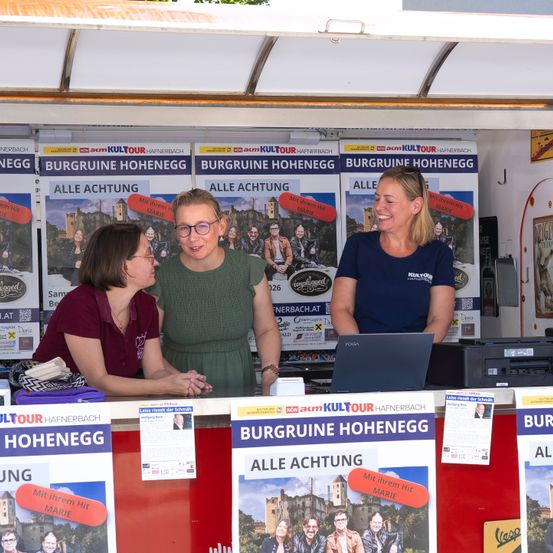 Drei Frauen stehen hinter einem Tresen in einem Stand, lächeln und schauen auf einen Laptop. Der Stand ist mit Plakaten gefüllt, die Burgruine Hohenegg bewerben.