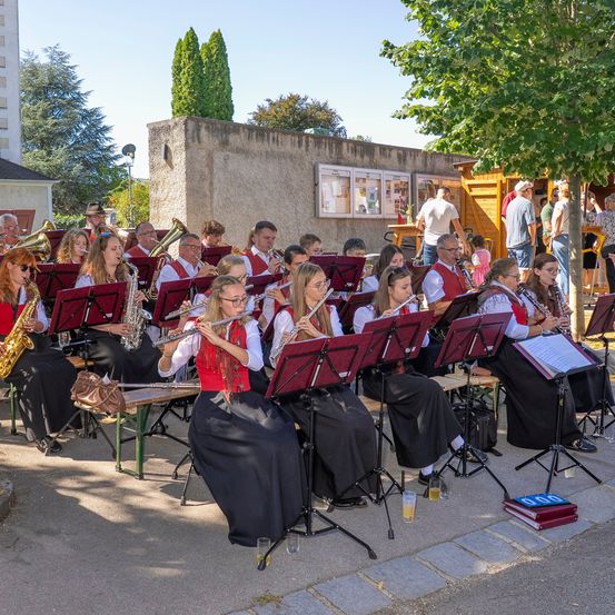 Eine Gruppe von Musikern in traditioneller Kleidung spielt an einem sonnigen Tag im Freien. Sie sitzen auf Bänken und spielen verschiedene Instrumente wie Flöten, Saxophone und Trompeten.