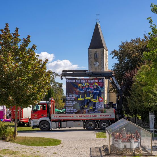 Ein Feuerwehrwagen mit einer großen Werbung steht vor einer Kirche mit Turm und Uhr unter einem klaren Himmel.
