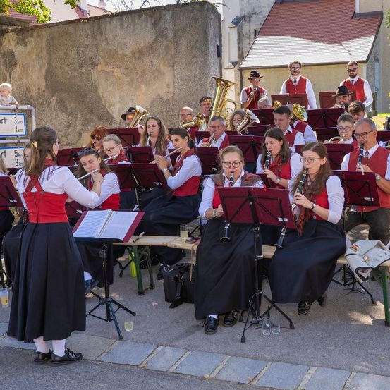 Eine Band spielt im Freien, wobei die Musiker verschiedene Instrumente spielen. Die Gruppe ist in traditioneller Kleidung gekleidet und sitzt auf Bänken. Die Umgebung scheint ein öffentliches Ereignis zu sein.