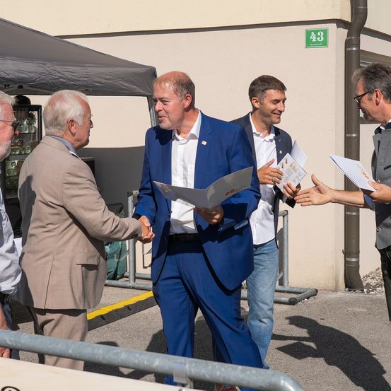 Ein Mann in einem blauen Anzug gibt einem anderen Mann in einem braunen Anzug die Hand. Sie stehen vor einem Gebäude.