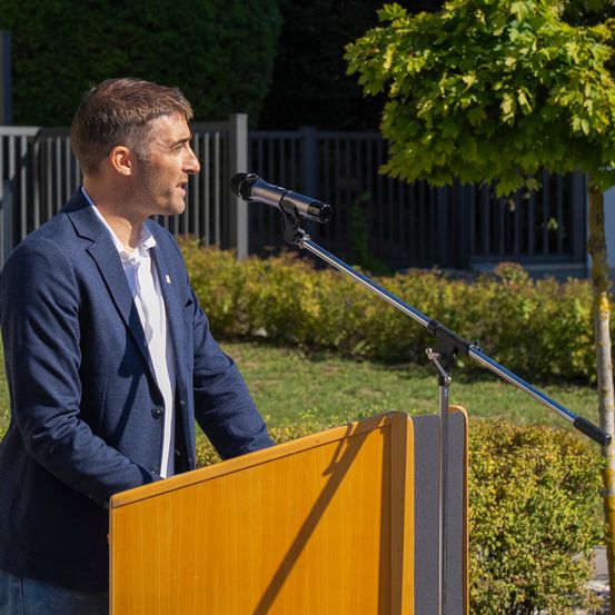 Ein Mann in einem blauen Blazer spricht in ein Mikrofon an einem Rednerpult, mit einem Baum und Hecken im Hintergrund.