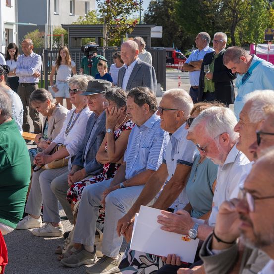 Eine Gruppe älterer Menschen sitzt auf Bänken in einer Außenumgebung, einige mit Brille und halten Papiere. Hinter ihnen stehen andere und schauen zu, wobei ein Feuerwehrwagen im Hintergrund zu sehen ist.