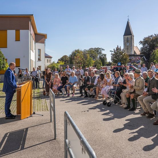 Ein Mann in einem blauen Anzug steht an einem Rednerpult und spricht eine Menschenmenge an, die auf der Straße vor einem modernen Gebäude sitzt. Im Hintergrund sind Bäume und ein Glockenturm zu sehen.