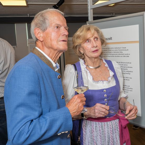 Ein älteres Paar steht eng beieinander, der Mann hält ein Weinglas, die Frau eine Brille. Sie stehen vor einem Plakat mit deutschem Text.