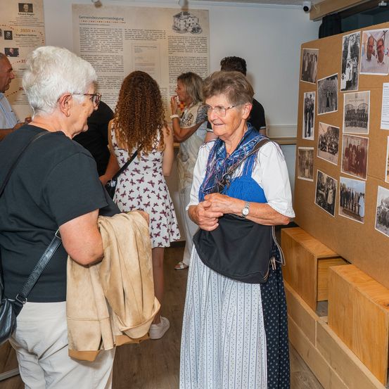 Eine Gruppe älterer Frauen steht in einem Raum, eine von ihnen trägt ein traditionelles Kleid und schaut auf eine Pinnwand mit alten Fotos.