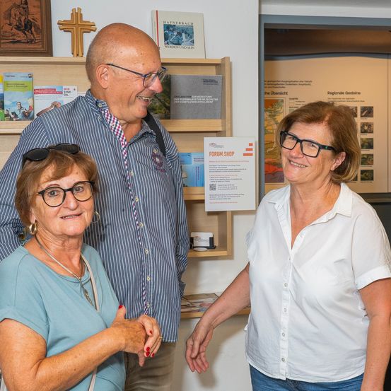 Drei Menschen stehen in einem Raum, lächeln und schauen in die Kamera. Der Mann trägt eine Brille und einen Rucksack. Die Frau links trägt eine Halskette und Ohrringe. Hinter ihnen steht ein Regal mit Büchern und einem Kreuz.
