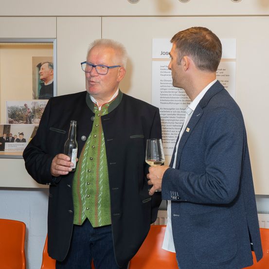 Zwei Männer in formeller Kleidung stehen in einem Büro, einer hält eine Flasche und der andere ein Weinglas. Hinter ihnen zeigt eine Wand ein Bild und Text.