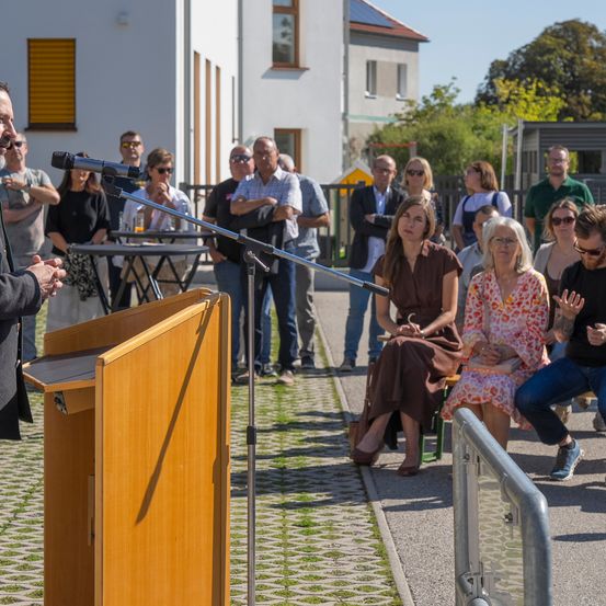 Ein Mann spricht an einem Podium, während ein vielfältiges Publikum aufmerksam zuhört. Der Ort ist im Freien mit Gebäuden und Grünpflanzen im Hintergrund.