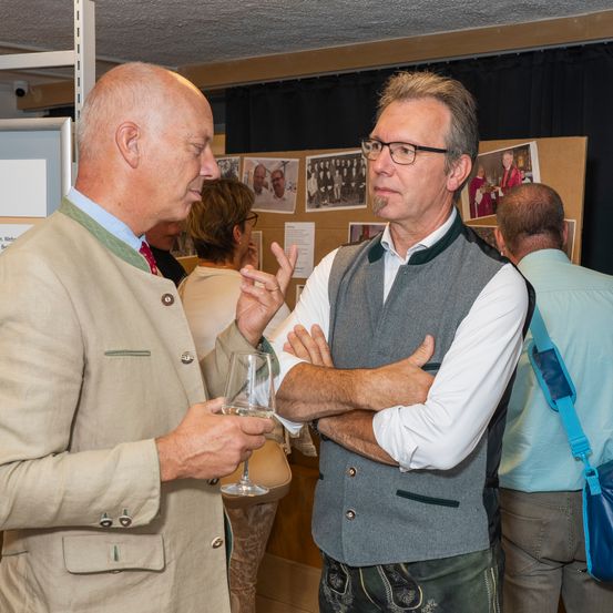 Zwei Männer in traditioneller Kleidung unterhalten sich bei einer Veranstaltung, einer hält ein Weinglas, während andere im Hintergrund herumlaufen.