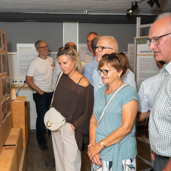 Eine Gruppe von Menschen steht in einem Raum mit Holzkisten und Postern an der Wand. Sie schauen sich die Poster an und scheinen etwas zu besprechen.