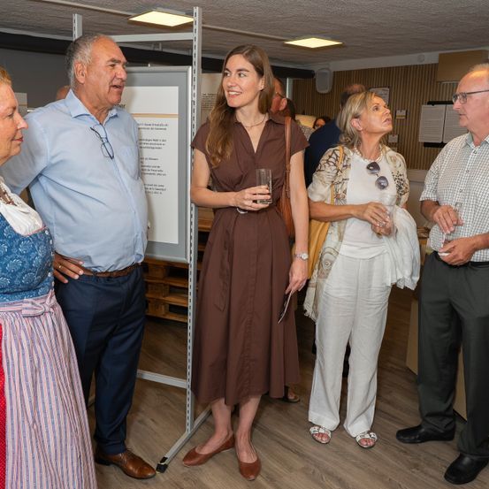 Eine Gruppe von Menschen steht in einem Raum, wahrscheinlich bei einer Ausstellung. Eine Frau in einem braunen Kleid hält ein Glas. Hinter ihr steht ein Mann in einem blauen Hemd, der lächelt.