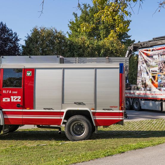 Ein roter und silberner Feuerwehrwagen steht am Straßenrand, mit einem Banner auf dem dahinterstehenden Lkw.