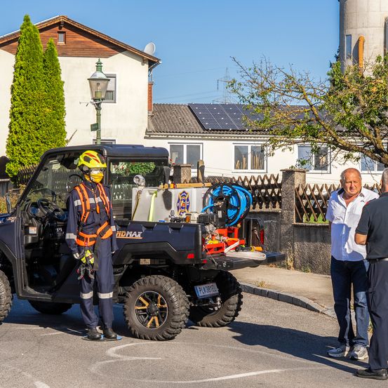 Ein Feuerwehrmann in voller Ausrüstung steht neben einem Versorgungsfahrzeug mit Ausrüstung, während zwei Männer in Freizeitkleidung in der Nähe stehen. Hinter ihnen steht ein Haus mit Solarpanelen auf dem Dach und einem Baum.