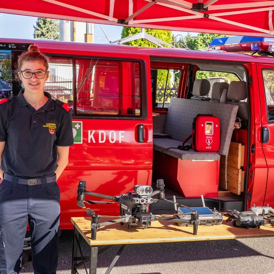 Eine Frau in Uniform steht neben einem roten Van mit den Buchstaben KDOF auf der Seite. Die Tür des Vans ist geöffnet und zeigt einen Tisch mit verschiedenen Drohnen darauf.