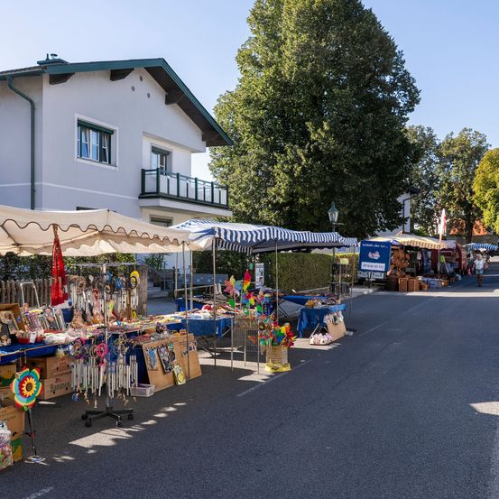 Ein Straßenmarkt mit verschiedenen Ständen unter bunten Sonnenschirmen, die Artikel wie Windspiele und Spielzeug verkaufen, mit einem Haus und Bäumen im Hintergrund.