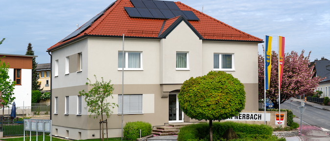 Ein zweistöckiges Haus mit Solarmodulen auf einem roten Dach steht an einer Straßenecke, umgeben von Grünflächen und Flaggen.