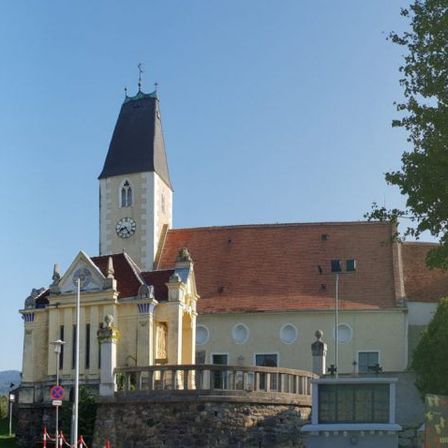 Eine Kirche mit Glockenturm steht prominent in einer Stadt, umgeben von Bäumen und Grün, mit einer Steinmauer und Geländern davor.