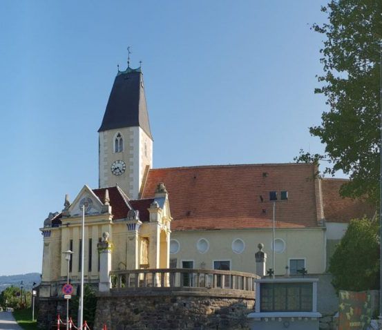Eine Kirche mit Glockenturm steht prominent in einer Stadt, umgeben von Bäumen und Grün, mit einer Steinmauer und Geländern davor.