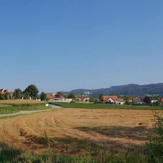 Ein breites, geerntetes Feld mit einem entfernten Dorf und Hügeln unter einem klaren blauen Himmel.