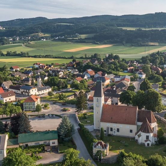 Luftaufnahme eines Dorfes, umgeben von grünen Feldern und Hügeln. Es zeigt eine Kirche mit einem hohen Turm, zahlreiche Häuser und üppige Bäume. Im Hintergrund befindet sich eine landschaftliche Szene mit sanften Hügeln.