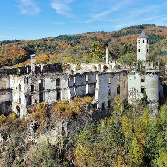 Auf einem Hügel umgeben die Ruinen einer alten Burg farbenfrohe Herbstlandschaft. Die Burg hat einen Turm und mehrere Gebäude, alle in einem Zustand des Verfalls.