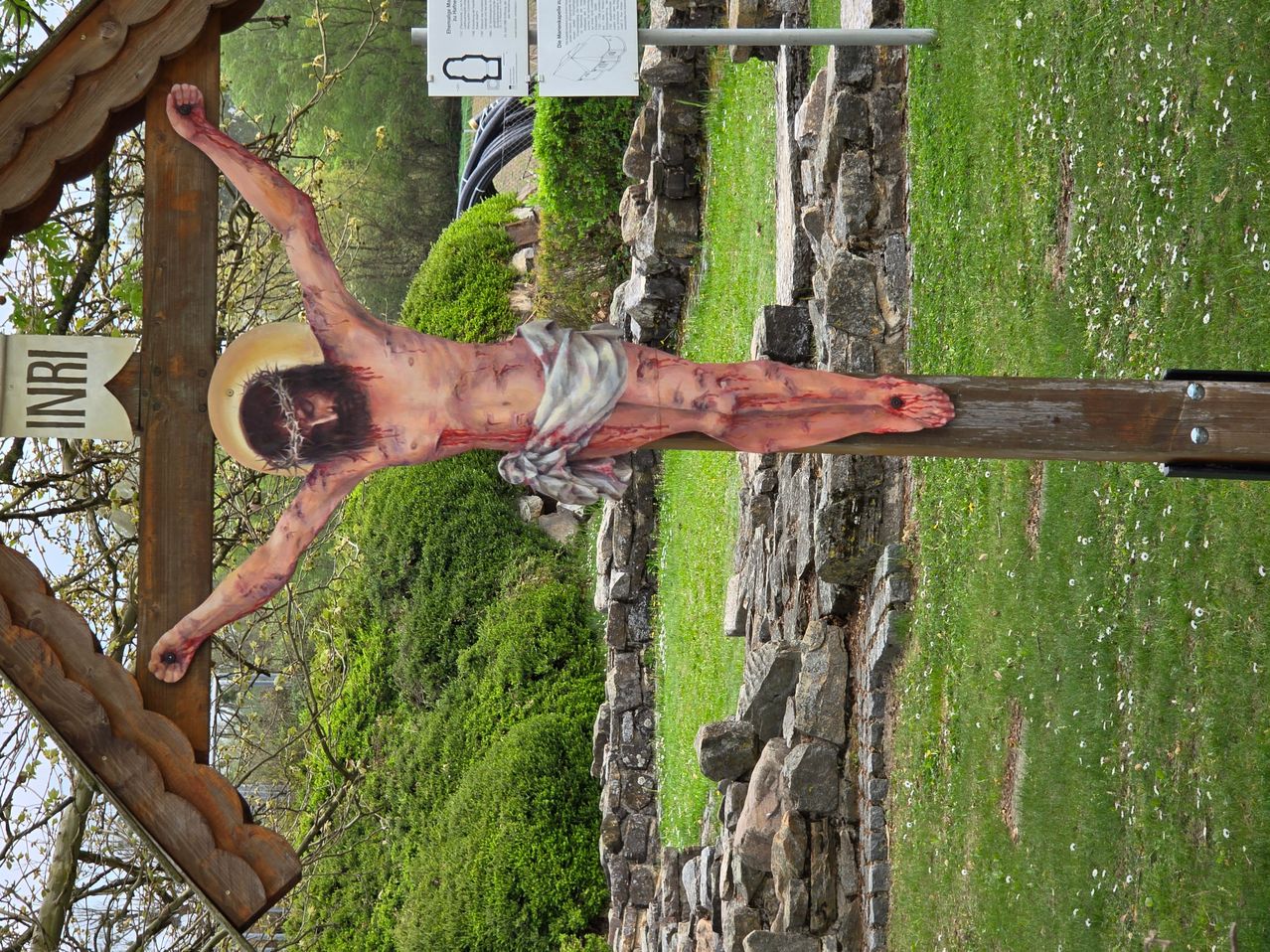 Ein Kruzifix mit einer detaillierten Darstellung von Jesus Christus, der ans Kreuz genagelt ist, vor dem Hintergrund einer Steinmauer und saftigem grünem Gras.