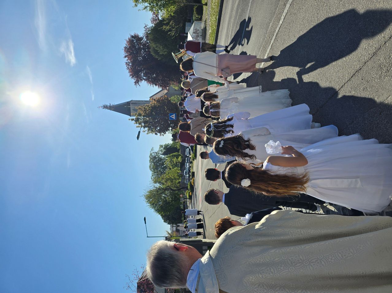 Eine Prozession mit vielen Menschen in weißen Kleidern und Anzügen, möglicherweise eine Hochzeit, findet auf einer Straße statt, mit einer Kirche im Hintergrund.